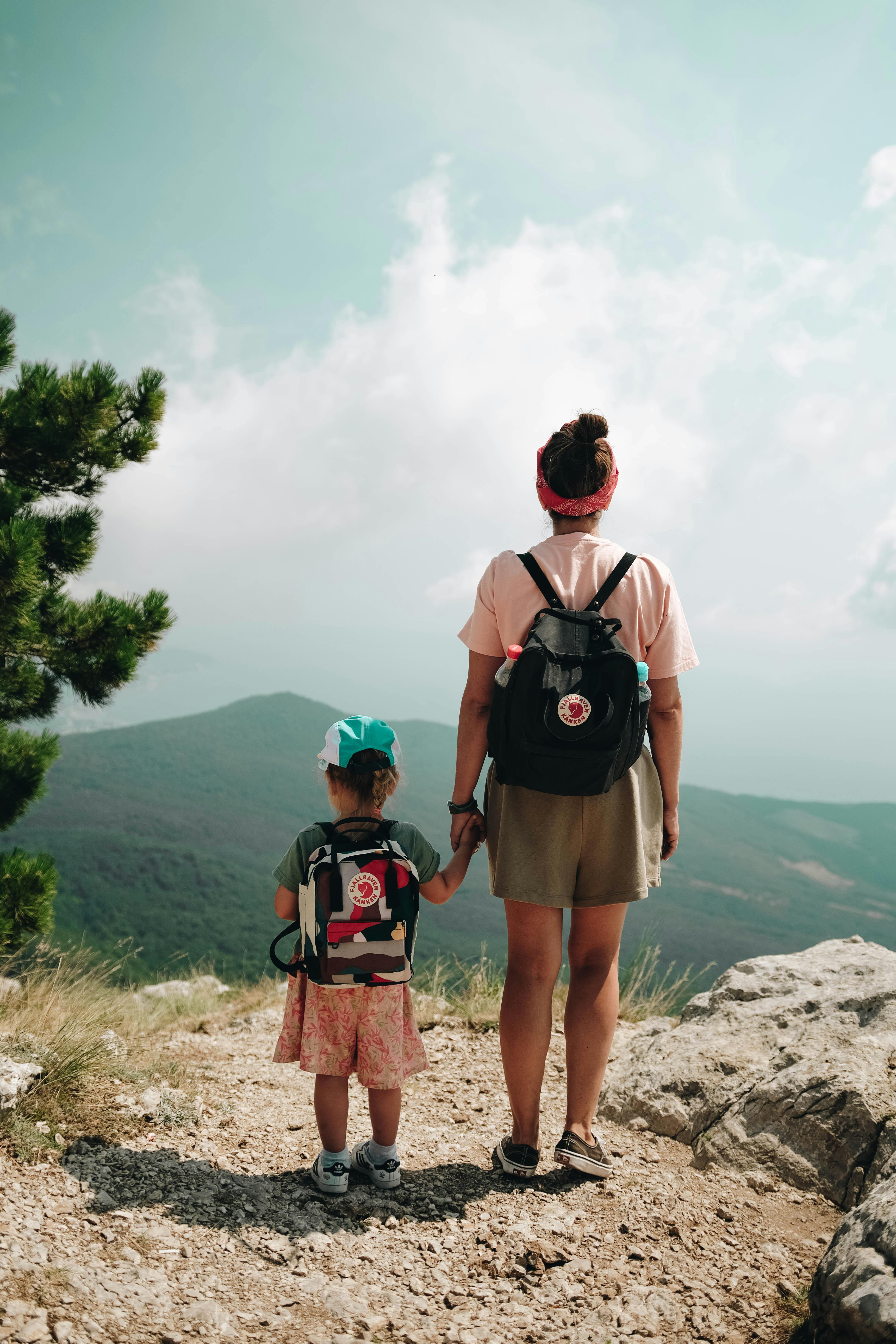 A woman and a child hiking on a mountain trail with backpacks, enjoying a scenic view.