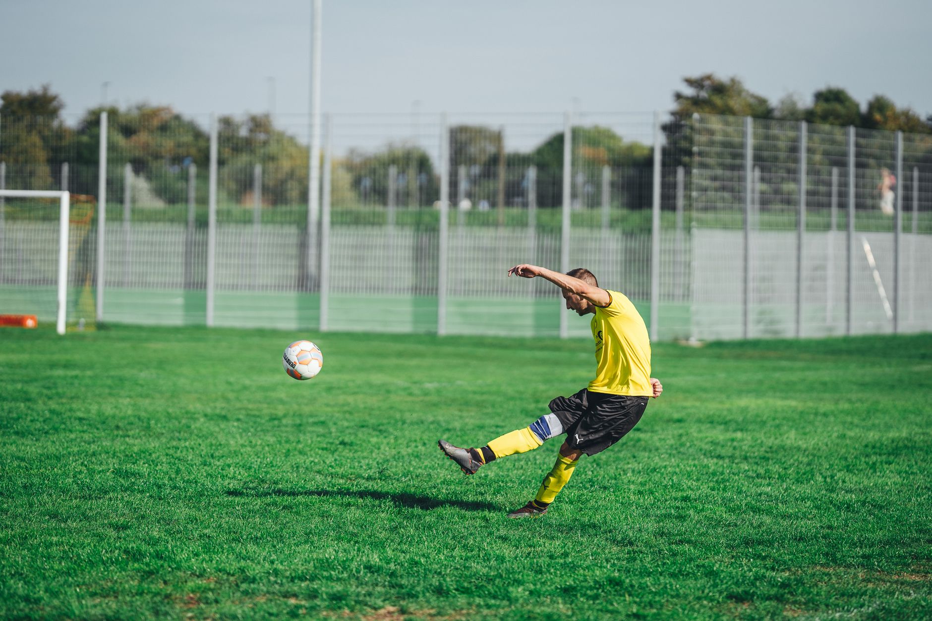 A male soccer player in a yellow jersey kicks a ball on a green outdoor field.