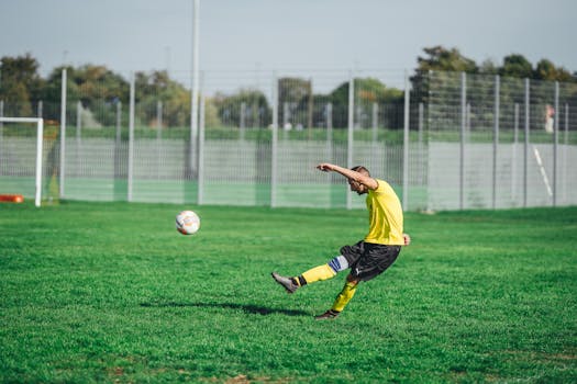 A male soccer player in a yellow jersey kicks a ball on a green outdoor field.