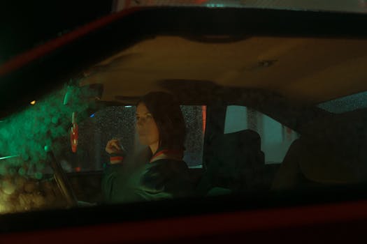 A woman sitting inside a parked car at night, gazing thoughtfully through the rain-speckled window.