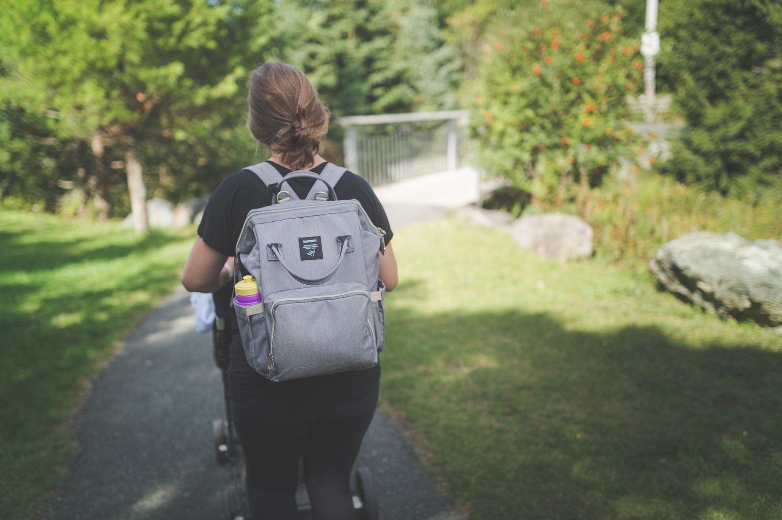 Parent wearing a gray backpack-style bag while walking a stroller through a park