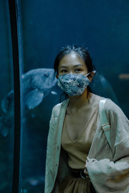 Woman in Beige Cardigan Wearing Face Mask while Standing Near an Aquarium