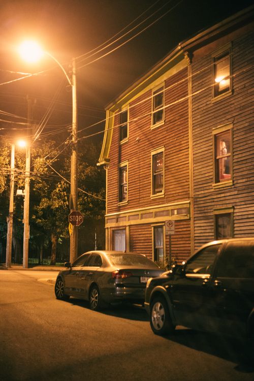 Free Urban nighttime scene with parked cars and a lit wooden building on a quiet street. Stock Photo