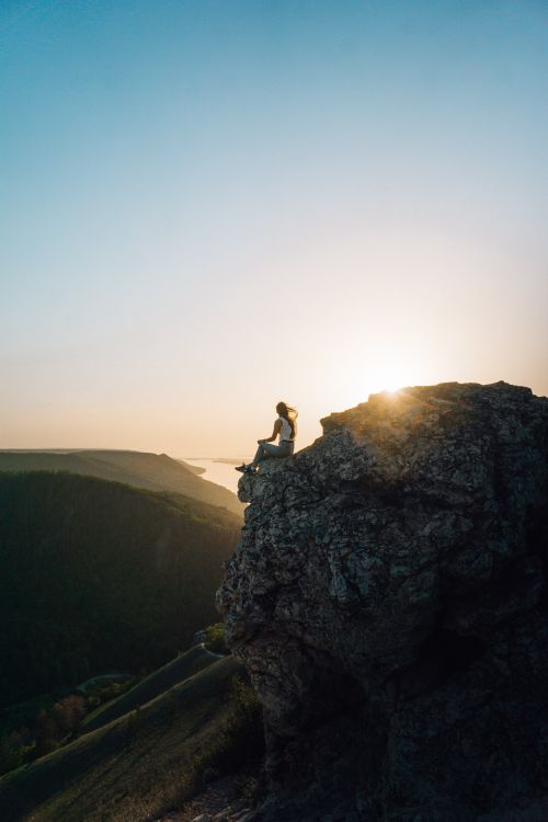 Free A woman sits on a cliff edge, enjoying the sunrise over a scenic mountain landscape. Stock Photo