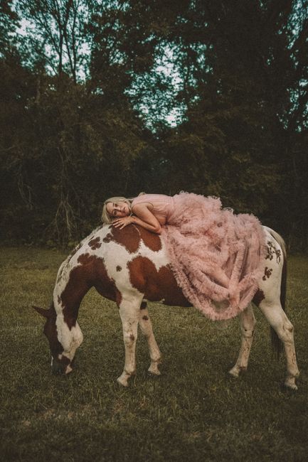 A woman in a pink gown lies on a spotted horse grazing in a serene outdoor setting.