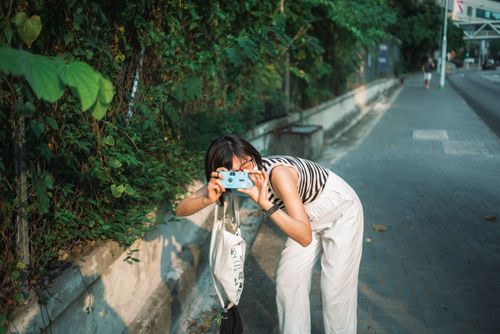 Free Casual outdoor shot of a woman taking pictures with a camera on a city sidewalk, surrounded by greenery. Stock Photo