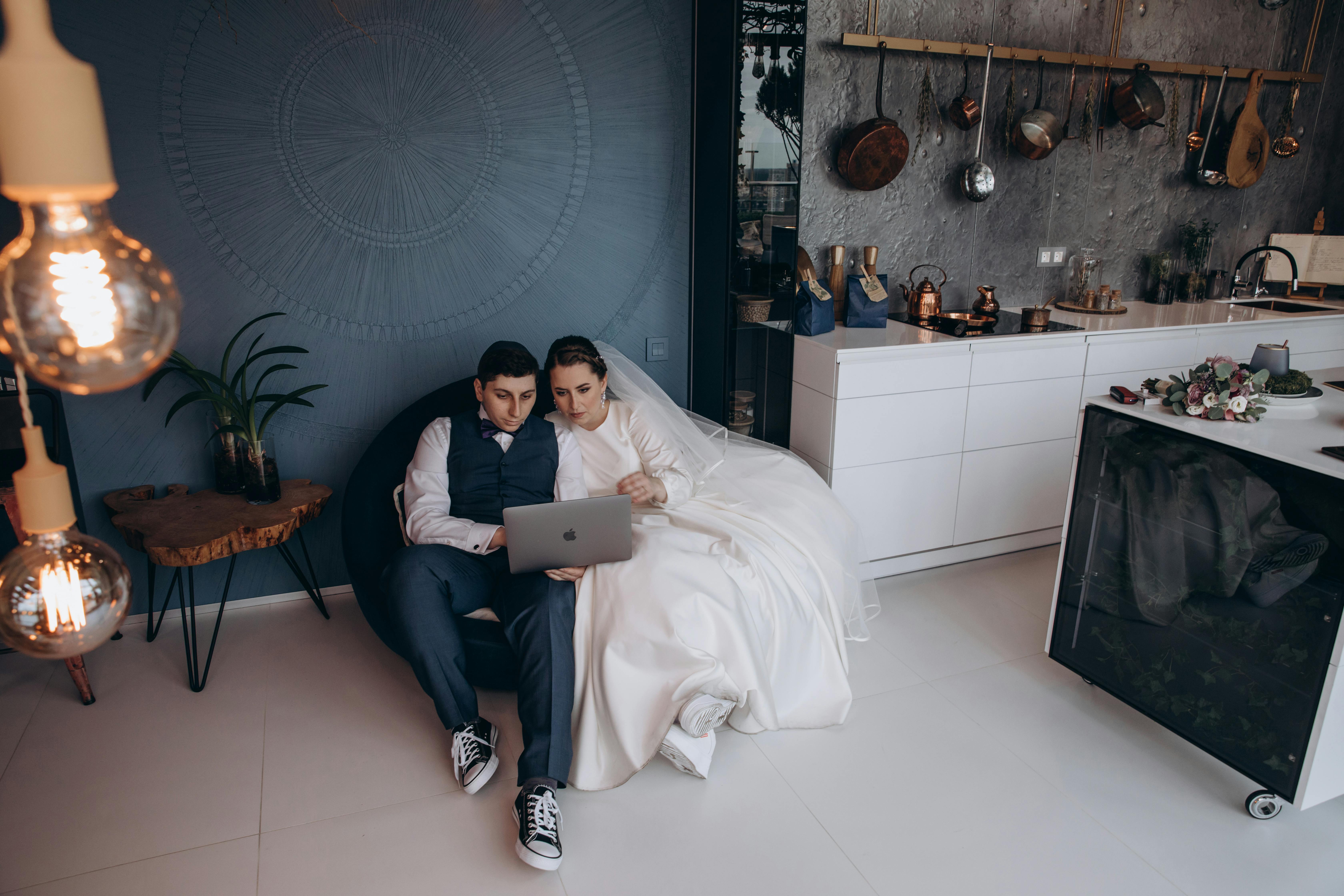 How Far in Advance Should You Book a Wedding DJ? - Bride and groom in kitchen reviewing wedding photos on a laptop, captured indoors.