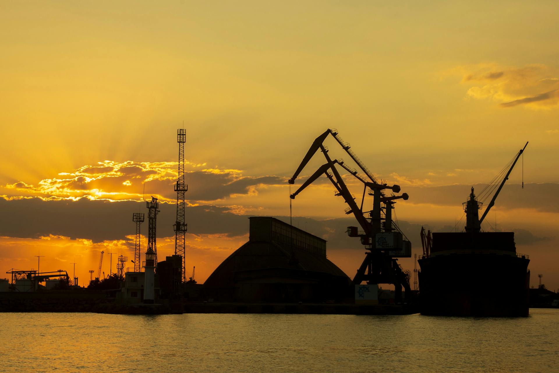 Silhouette of a port with cranes and ships during a vibrant golden sunset, creating a dramatic skyline.