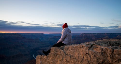 Person Wearing Gray Hooded Jacket and Black Pants Sitting on Mountain Cliff during Sunset