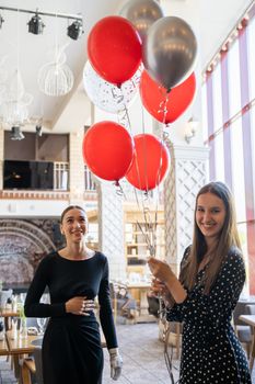 Two happy women enjoy a birthday celebration with red and silver balloons indoors.