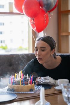 A woman blows out candles on a birthday cake with colorful balloons in the background.