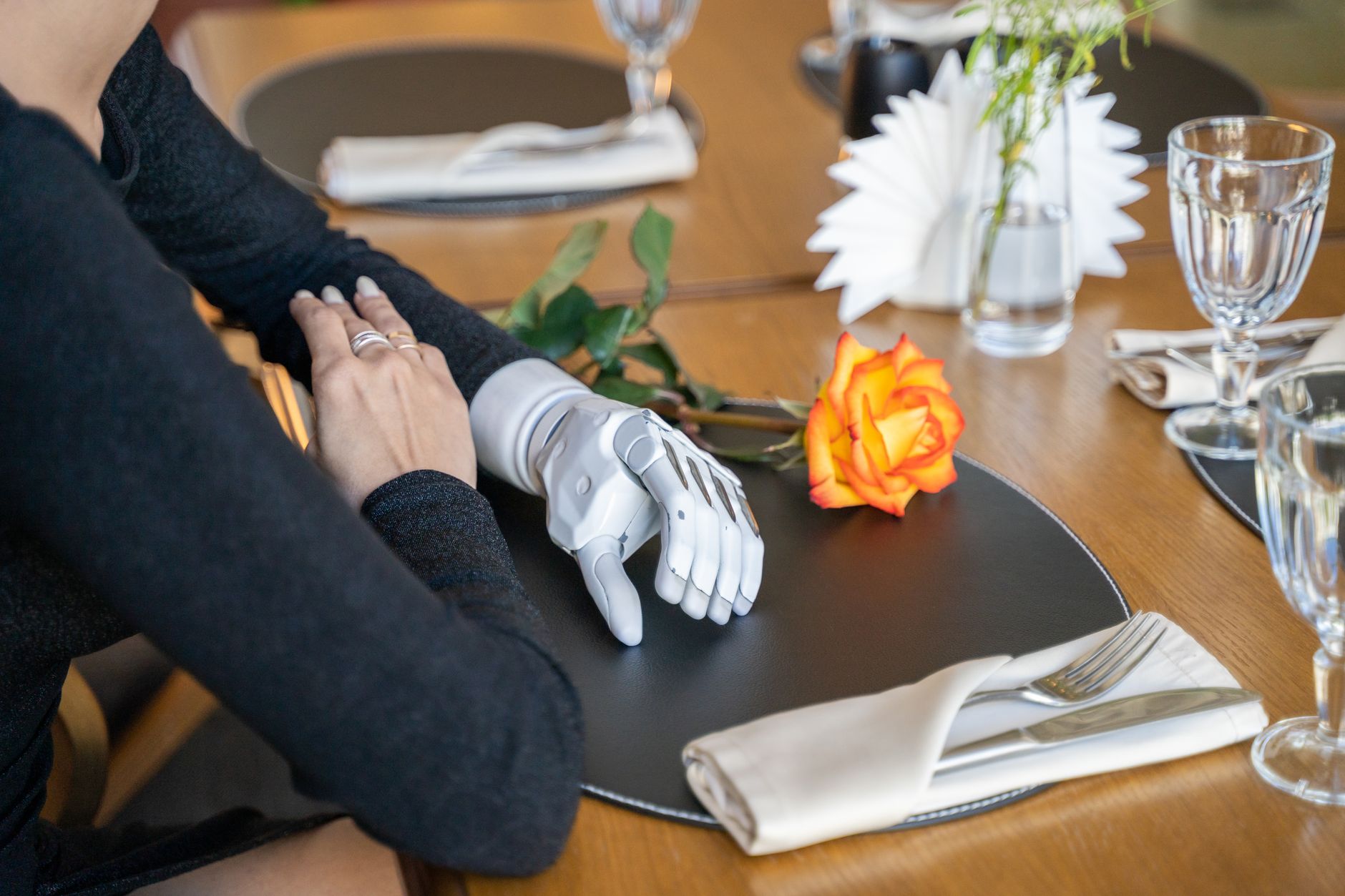 A dining setting featuring a robotic prosthetic arm and a single rose on a table.