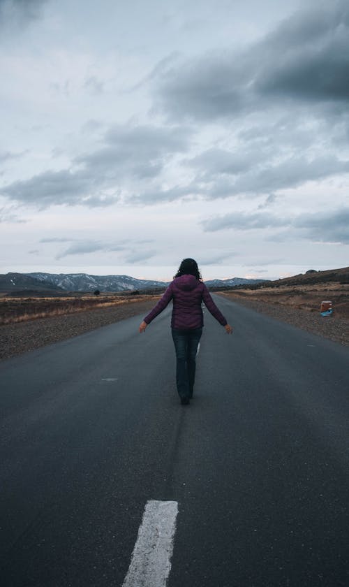 Free Back View of a Person Walking Alone in the Middle of the Road under the Cloudy Sky Stock Photo
