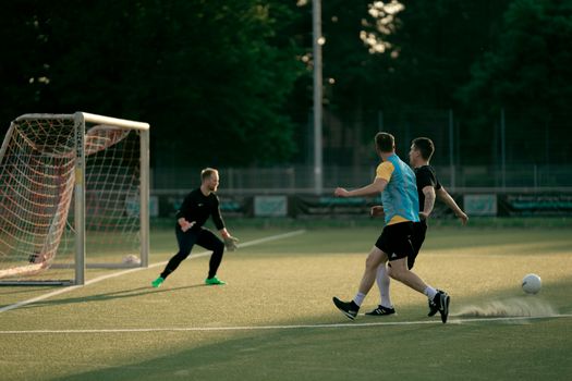Two soccer players and a goalie in action during an outdoor match at sunset.