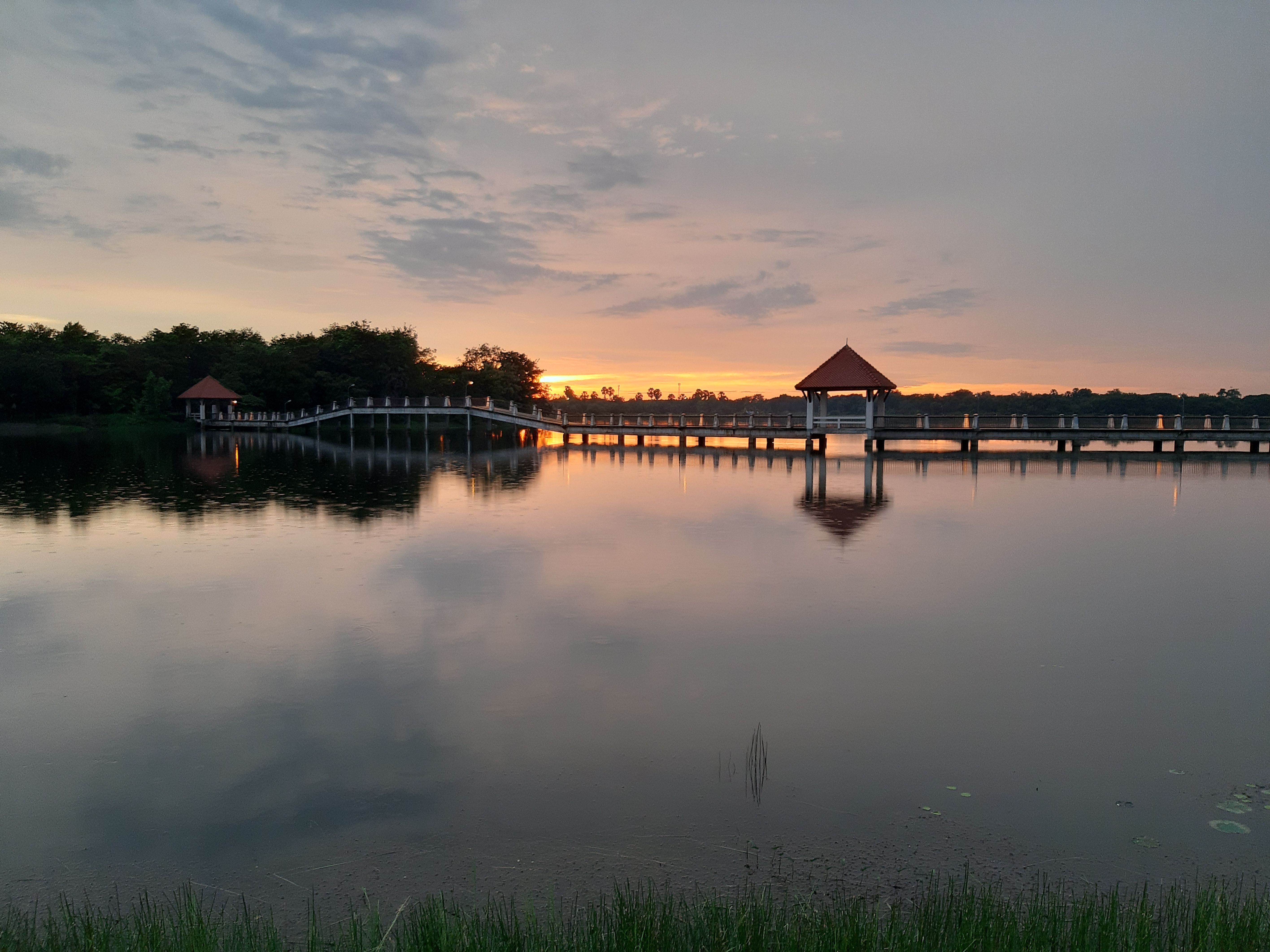 Serene sunset view over a bridge and gazebo reflecting in calm waters in Thailand.