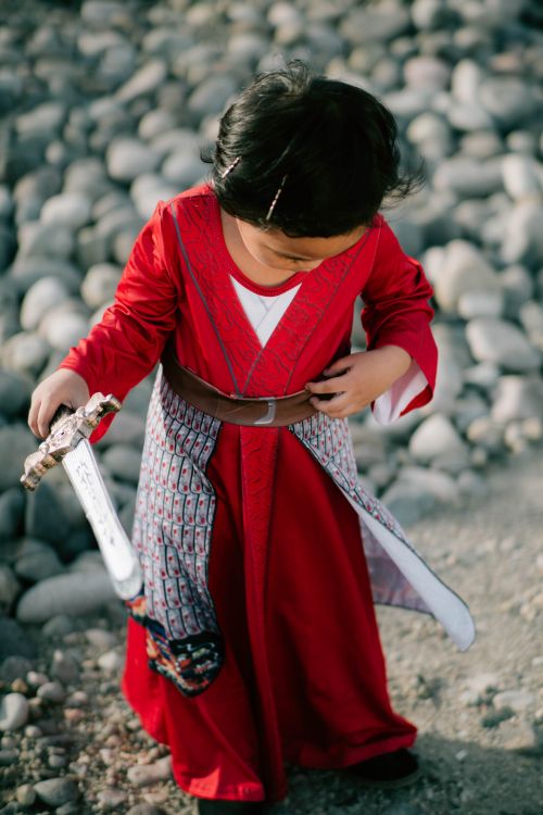 Free A young child in a red costume holds a toy sword while standing on a pebbled surface, looking down. Stock Photo