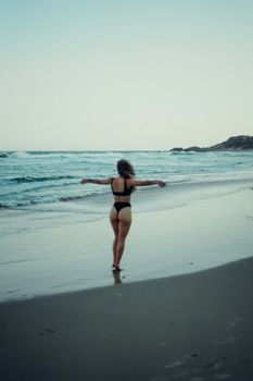 A woman walks along a sandy beach, wearing a black bikini with arms outstretched, enjoying the ocean waves.