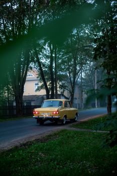 Classic yellow car driving through a peaceful, tree-lined road in the early evening.