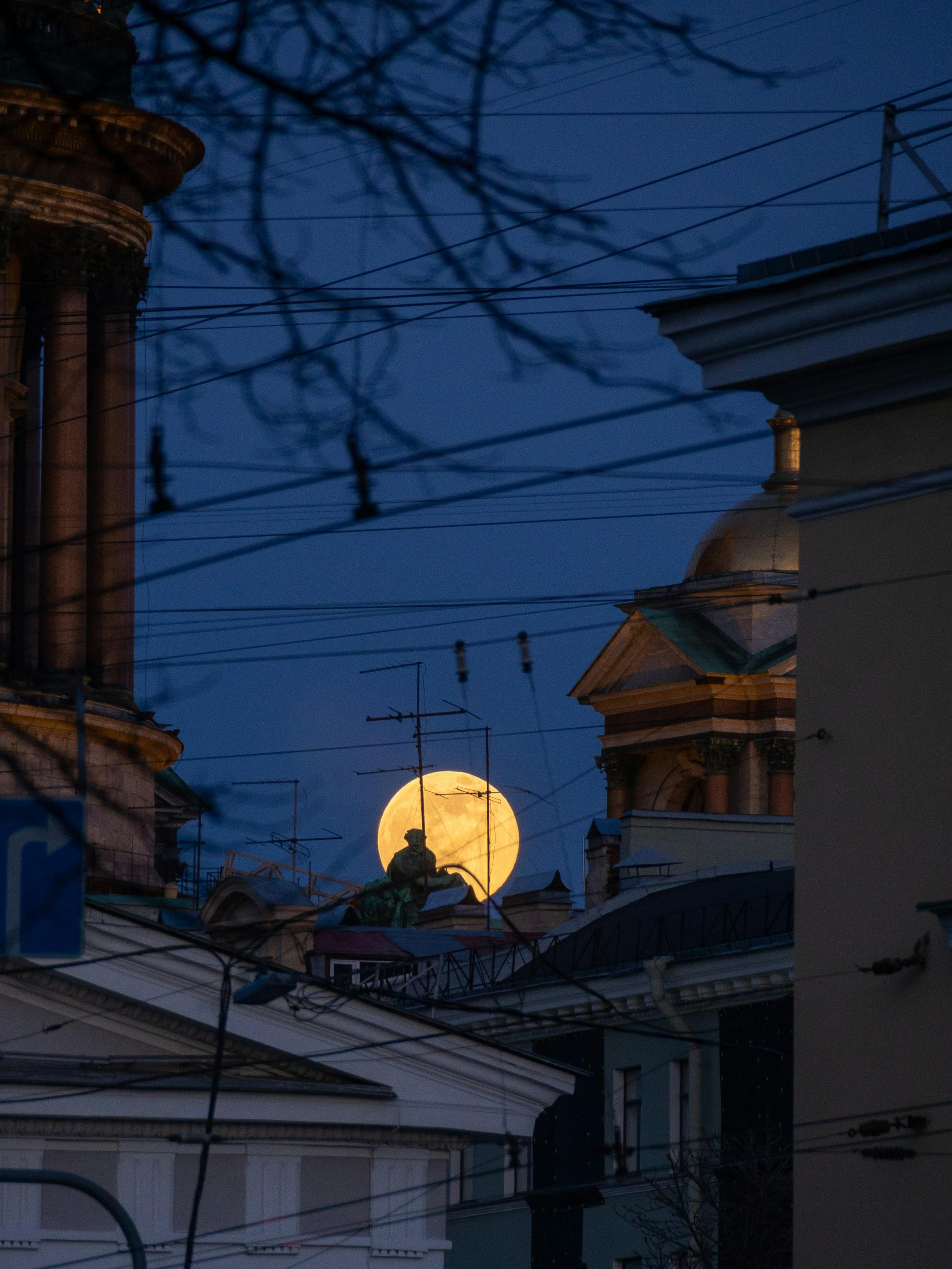 A full moon rises against an urban skyline with historic architecture, creating a dramatic night scene.