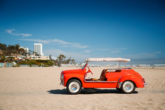 A classic red car parked on a sunny beach with a city skyline in the background.