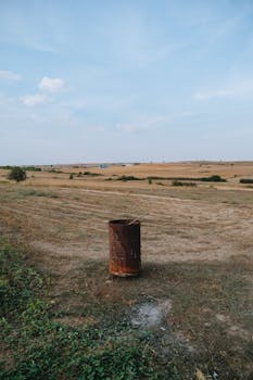 A rusty barrel in an expansive rural field under a clear blue sky.
