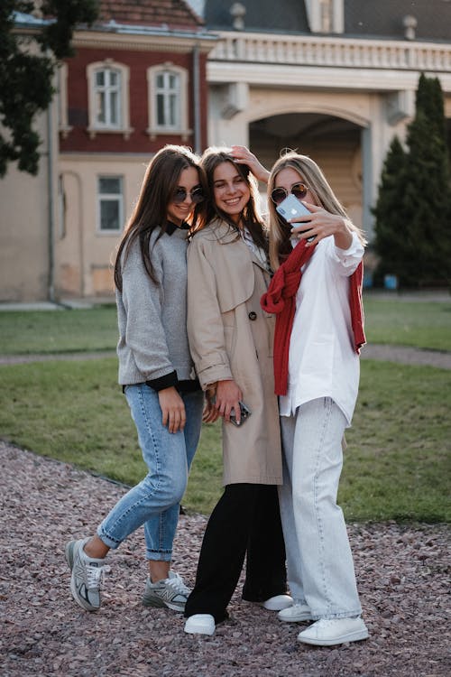 Full body of cheerful young female friends in casual outfits smiling and taking selfie on mobile phone while standing on street in city