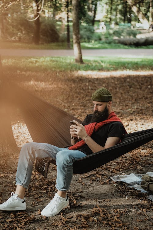 A Bearded Man Using His Cellphone while Sitting on a Hammock