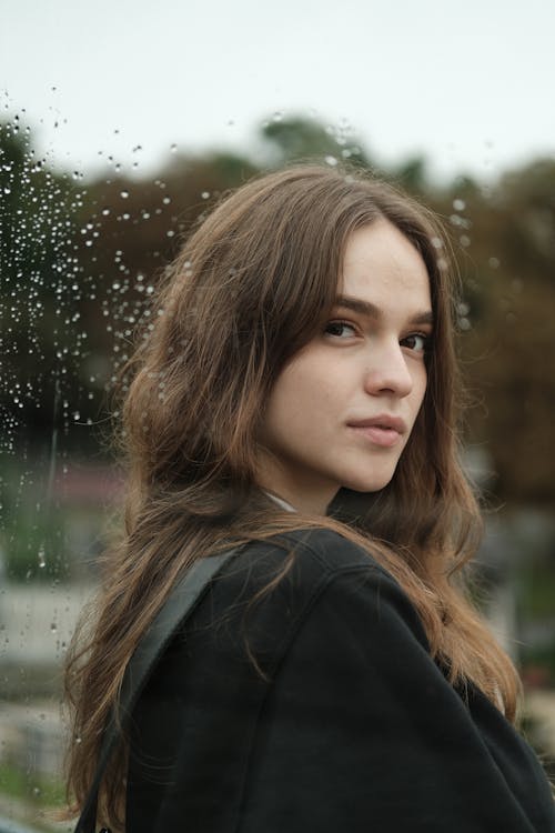 Portrait of young charming female brunette standing near glass wall with raindrops and looking at camera in sunlight
