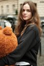 Young brunette female in black sweater with long wavy hair holding big brown toy bear and looking at camera on street