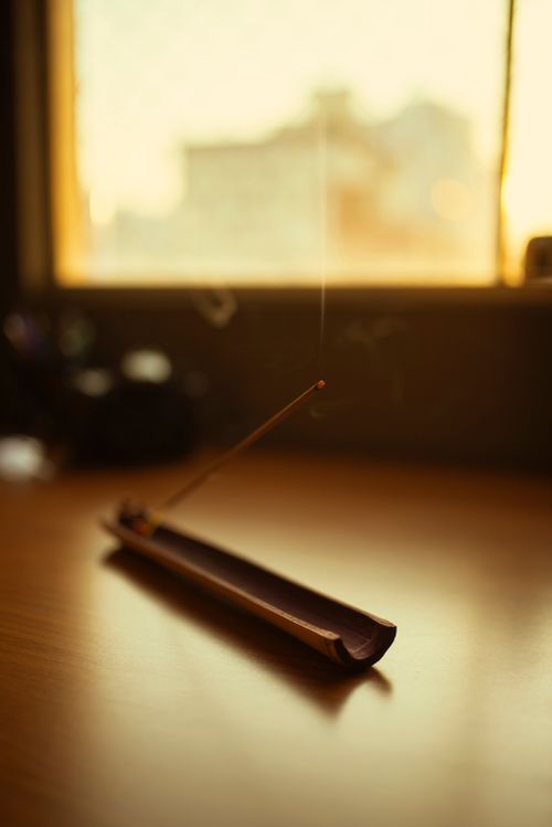 Free Incense stick burning on a wooden holder with smoke rising gently, creating a serene ambiance in a sunlit room. Stock Photo