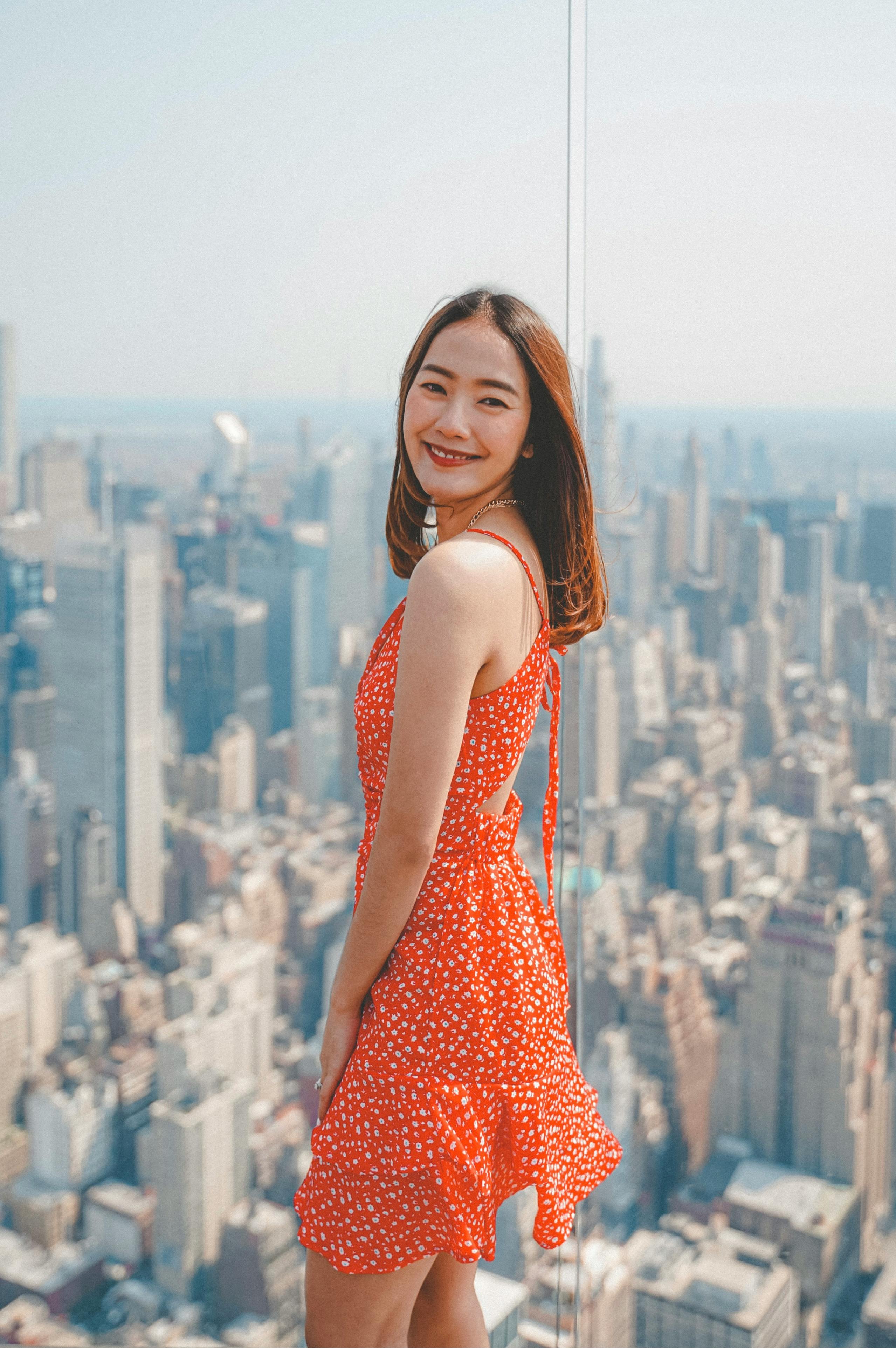 Asian woman posing on New York City skyscraper deck, smiling in a red dress.