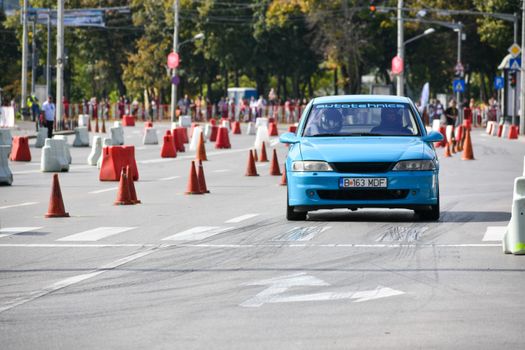 Blue car driving through an urban race course with traffic cones and onlookers.