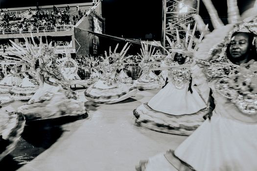 Elegant dancers in elaborate costumes perform in a grayscale parade scene at night.