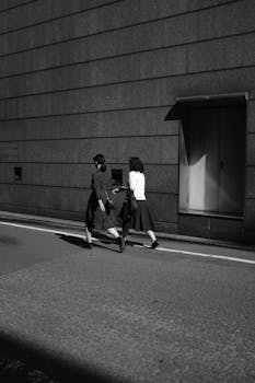 Two women walking by a shadowed wall in an urban Japanese street with a minimalistic vibe.