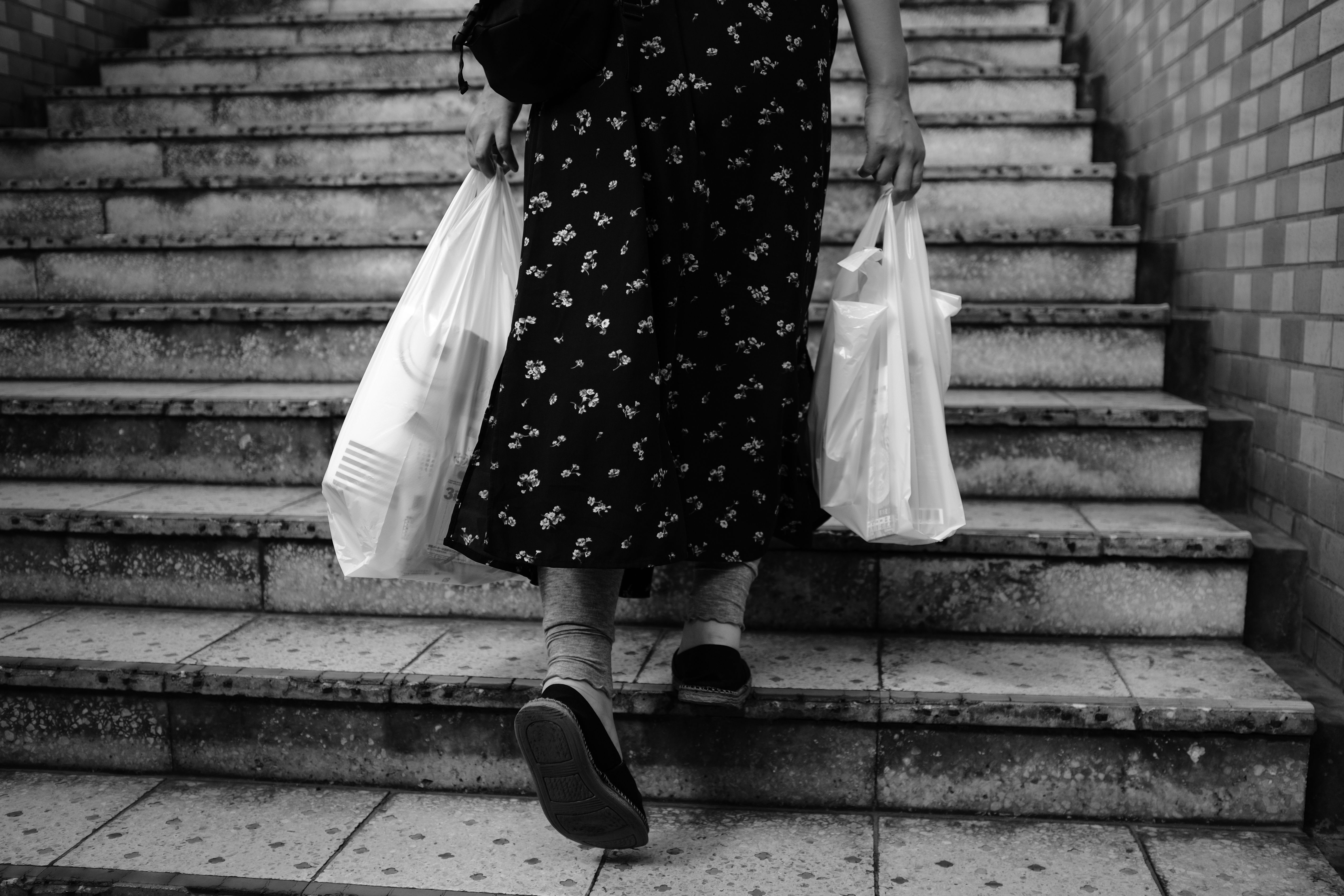 Black and white photo of a woman walking up stairs carrying plastic bags.