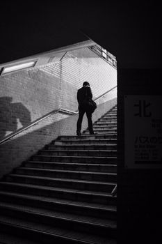 Black and white photograph of a man walking up a subway staircase in urban Japan.