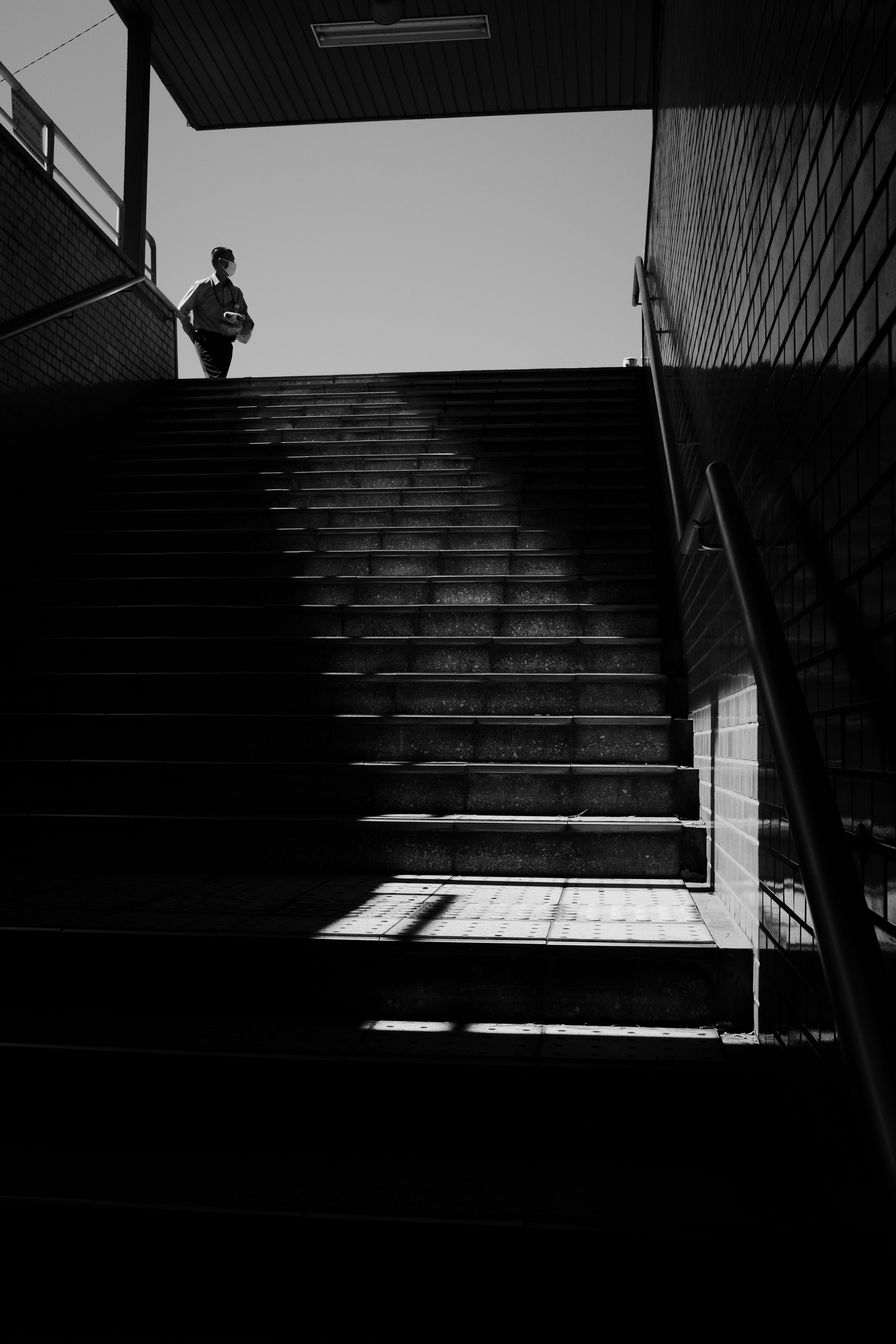 Moody black and white silhouette of a man standing at the top of shadowed stairs outdoors.