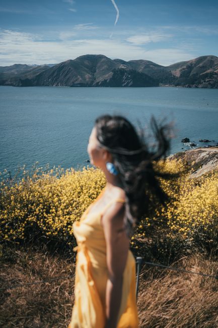 Woman in a yellow dress with flowing hair stands by a scenic ocean cliff with vibrant yellow flowers.