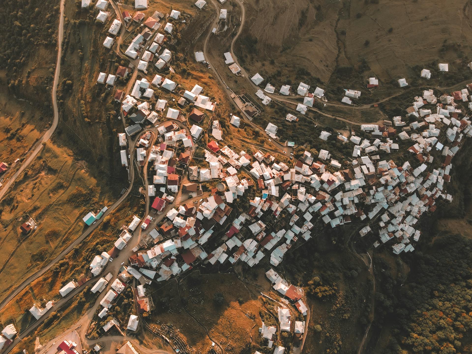 Aerial shot of a rural village with clustered houses under warm daylight, showcasing winding roads and lush surroundings.