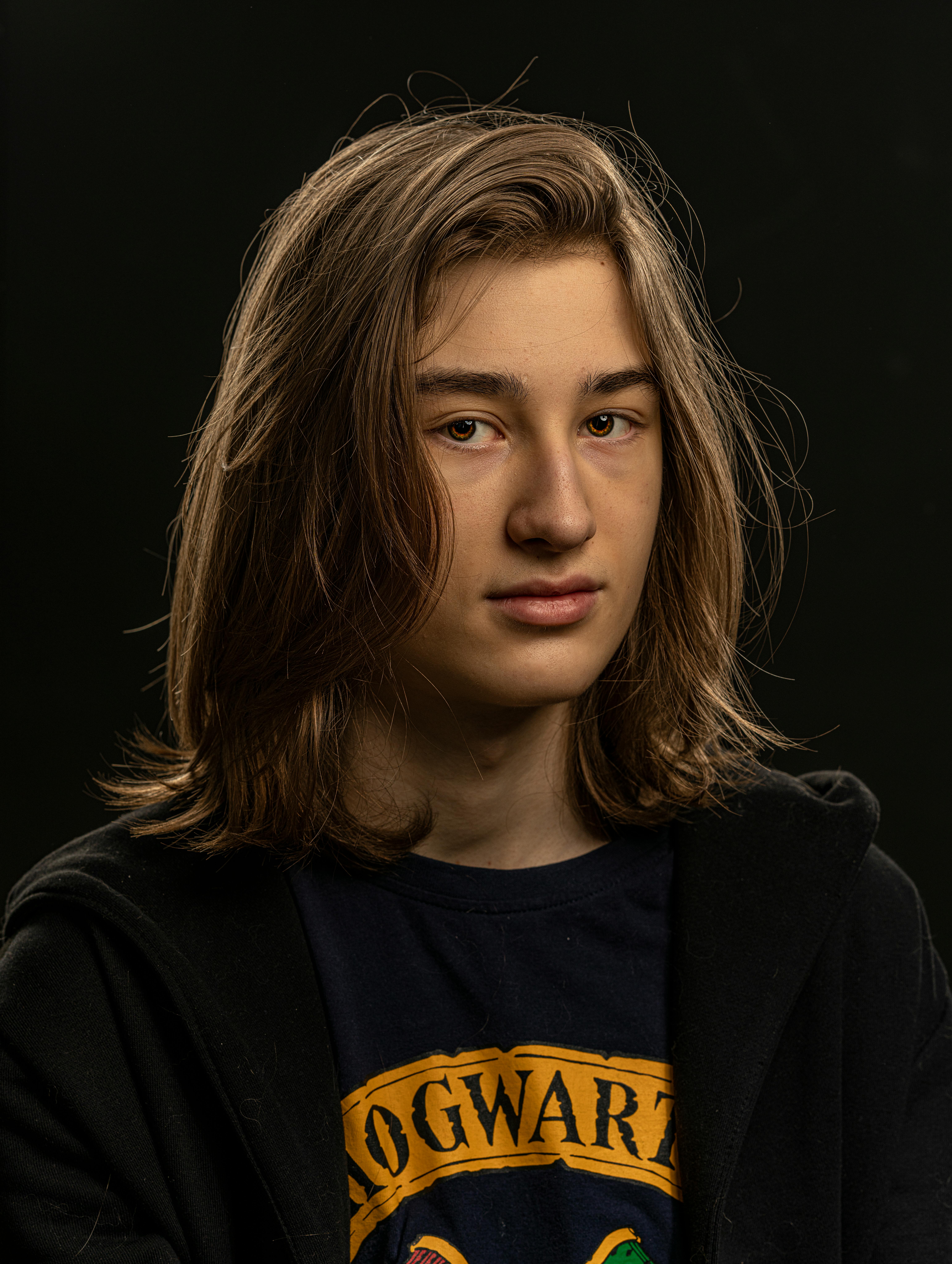 Close-up portrait of a teenage boy with long hair wearing a casual shirt and jacket in a studio setting.
