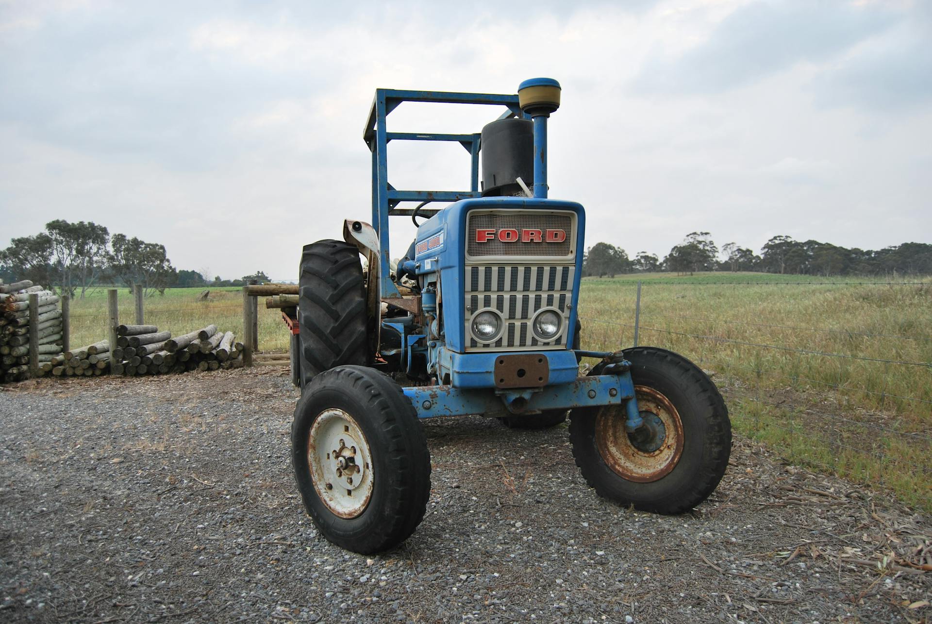 A classic blue Ford tractor parked on a gravel path in a rustic countryside farm environment.