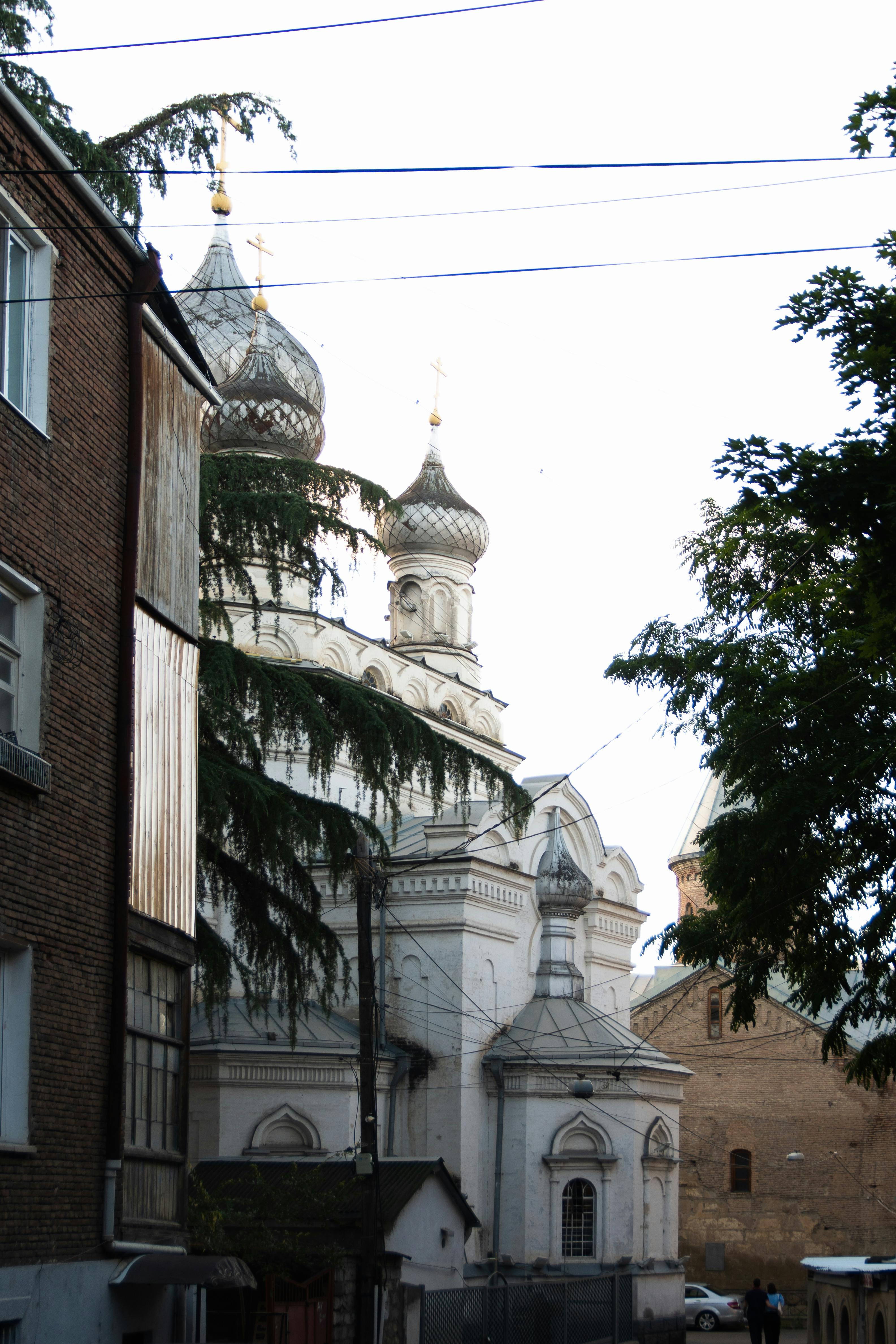 An ancient cathedral with domes in an urban city setting, featuring trees and adjacent buildings.