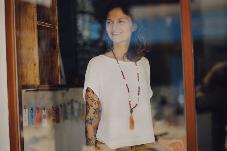 Woman In White Top With Arm Tattoos Behind A Glass