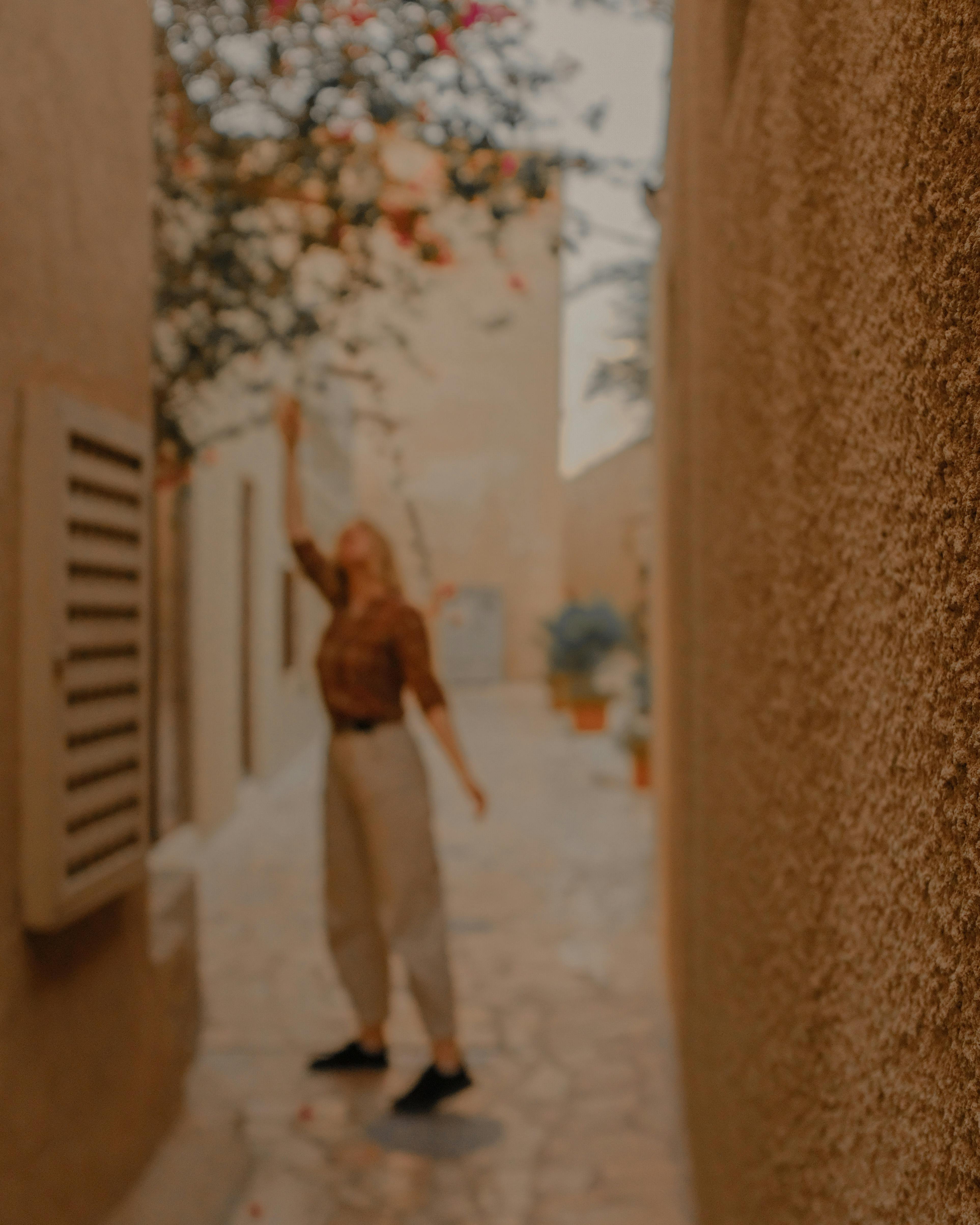 A woman reaches up to touch flowers in a tranquil, charming alleyway.