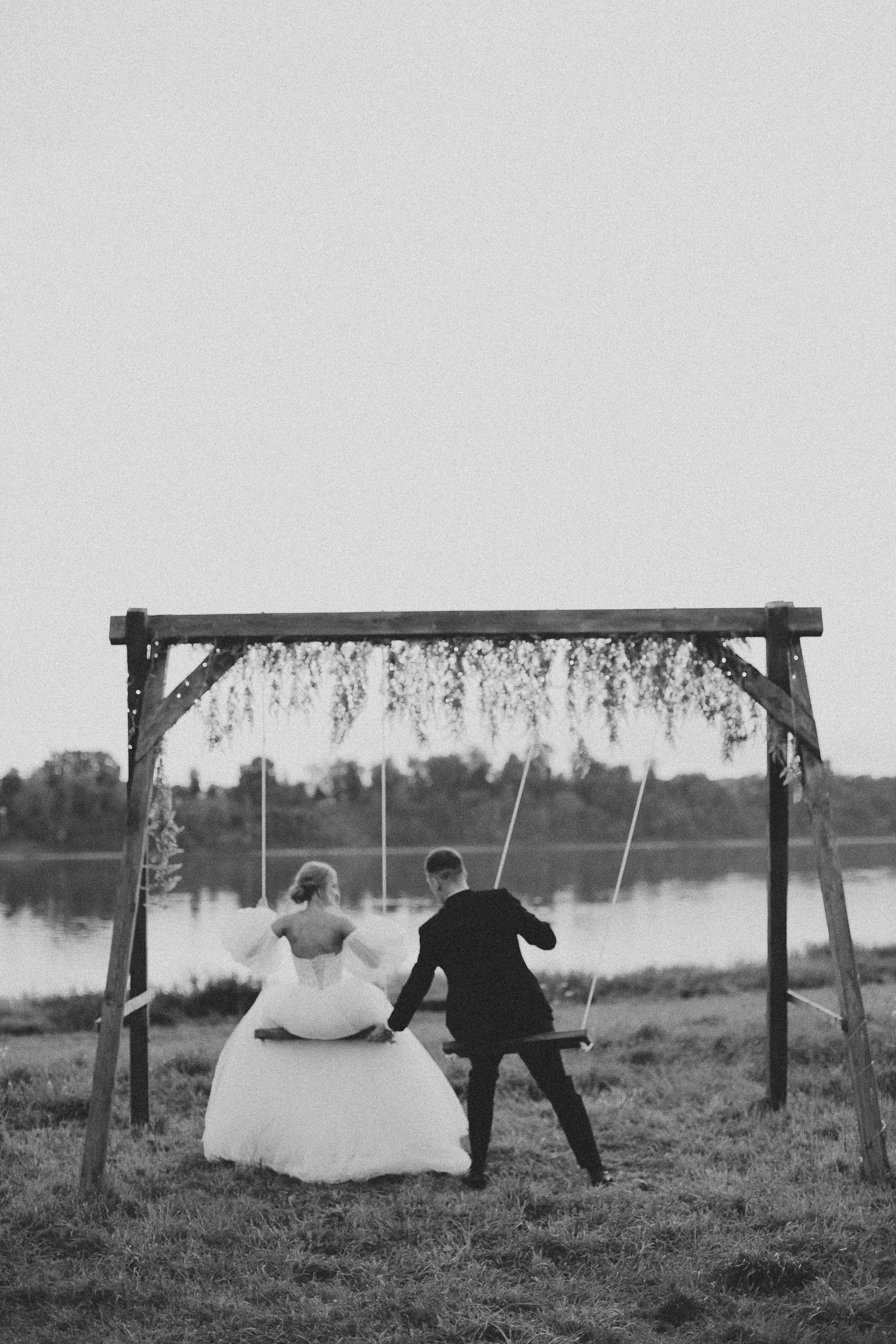 A romantic black and white photo of a wedding couple on a swing by a lake.