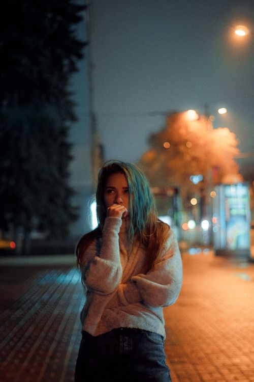 Free Portrait of a young woman in casual attire standing thoughtfully on a city street at night. Stock Photo