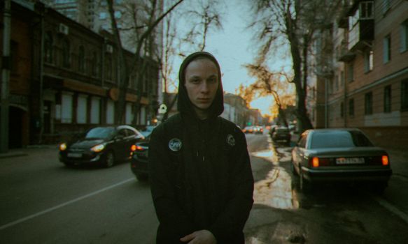 A young man in a hoodie stands on a dimly lit street, surrounded by city traffic.