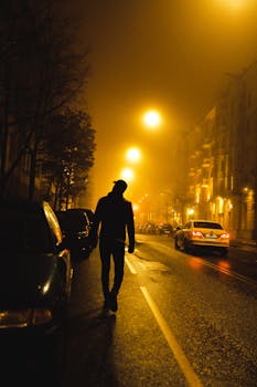 A mysterious silhouette walks along an illuminated city street, surrounded by mist and glowing streetlights at night.