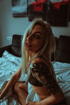 Blonde woman with tattoo sitting on a bed in a home interior.