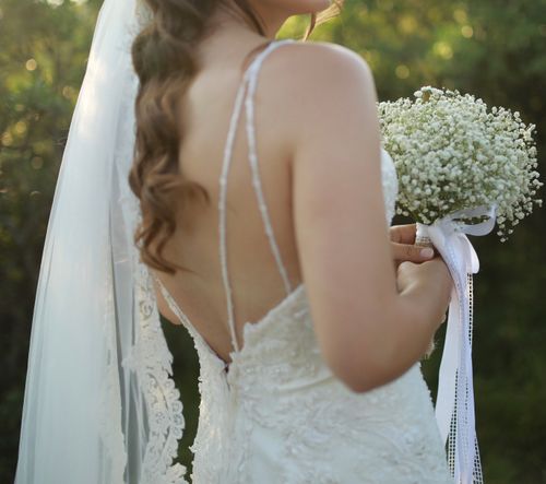 Free A serene back view of a bride in a white gown holding a bouquet outdoors. Stock Photo